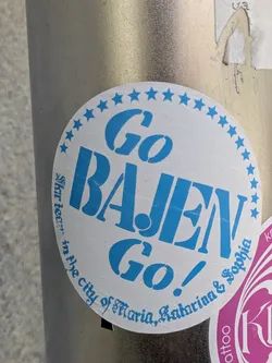 A round, light blue sticker on a metallic pole featuring the text "Go Bajen Go!" in large, bold letters. The sticker has a border of stars and includes the phrase "star team in the city of Maria, Katarina & Sophia".