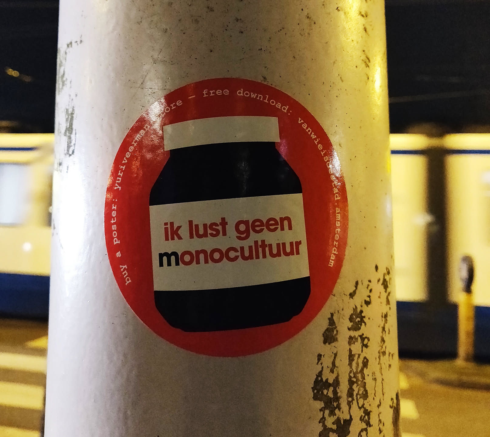 A round sticker on a pole depicting a jar with the text "ik lust geen monocultuur" which translates to "I don't like monoculture".  The sticker also mentions it's a free download from yuriveermagastore.com in Amsterdam.