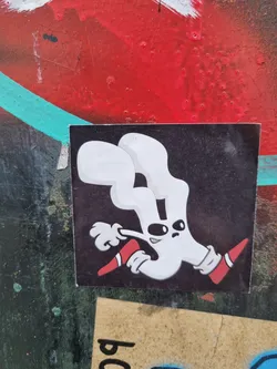 A black square sticker features a unique design of a white, almost hand-shaped creature. It has a simple, cartoonish style, with large eyes and red shoes or boots. The creature appears dynamic, as if in motion. The sticker has a matte finish, adding to its overall aesthetic.