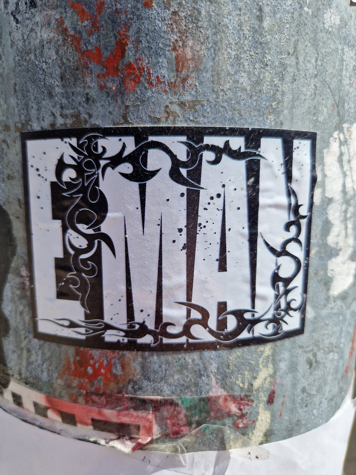A square sticker with a predominantly white background features a bold, stylized "MA" in a sans-serif font.  The letters are framed by a black tribal-esque design that wraps around the edges. The style is reminiscent of tattoo art or graffiti. The sticker shows some wear, suggesting it's been exposed to the elements.