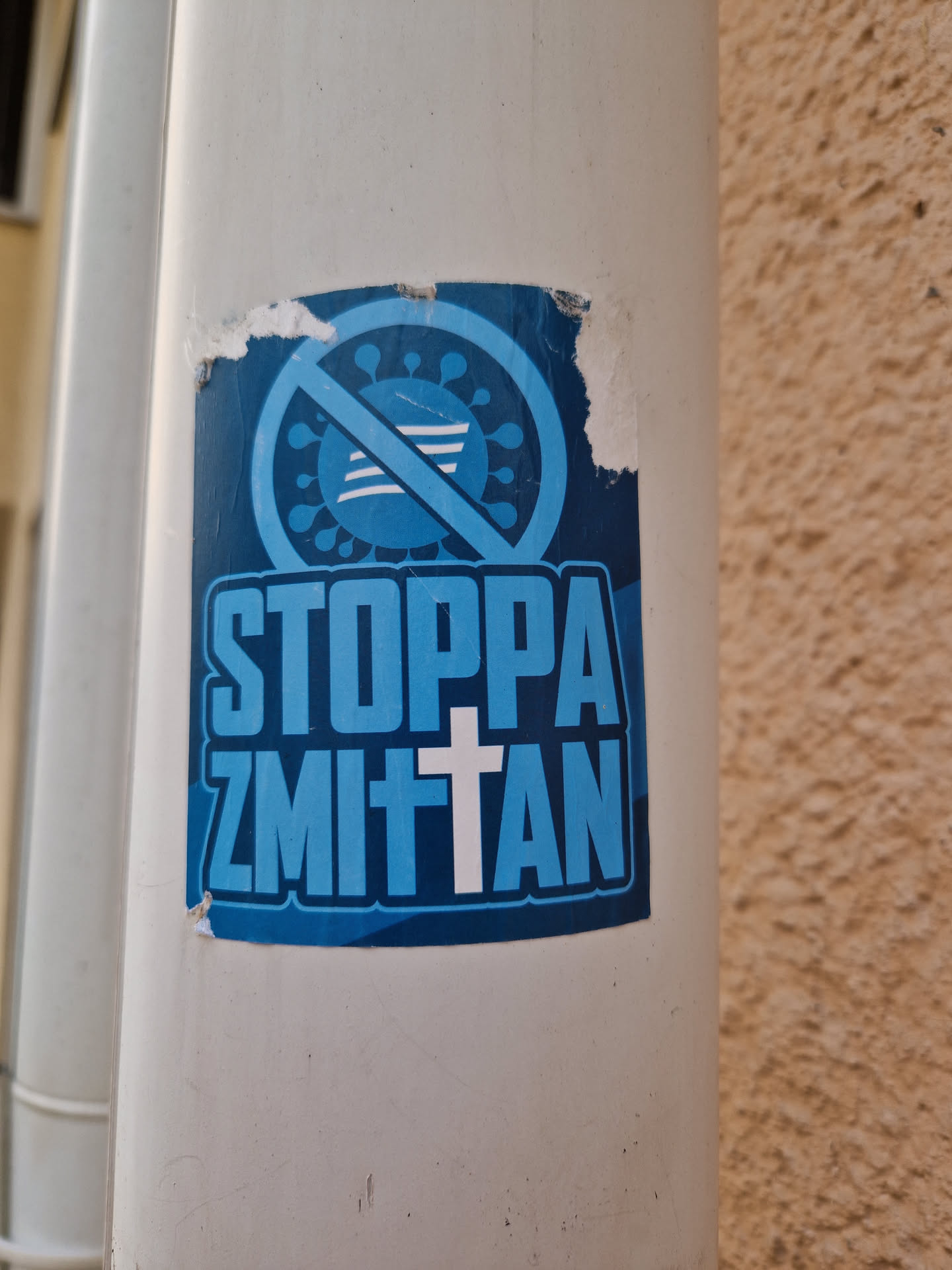 A blue sticker with a symbol of a virus with a cross on it and the text "Stoppa Zmitten". The sticker is slightly damaged.