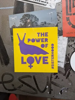 Street sticker A square adhesive sticker with a bold yellow background and purple graphic elements. In the center-left is the solid silhouette of a slug facing left with two prominent eye stalks. To the right of the slug's head and neck, bold sans-serif text reads 'THE POWER OF' stacked in three lines. Below the slug, the word 'LOVE' is printed in large block letters, with the letter 'O' designed as a female gender symbol (Venus symbol). Running vertically along the lower right border is the hashtag text '#SISTERHOOD'.