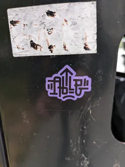 A small, round sticker with a purple background and black text. The text is stylized and includes an upward-pointing arrow.  The style is minimalist and graphic.