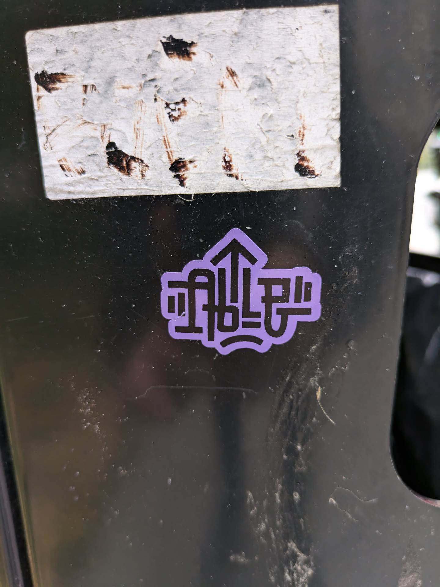 A small, round sticker with a purple background and black text. The text is stylized and includes an upward-pointing arrow.  The style is minimalist and graphic.