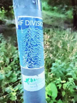 A sticker featuring the iconic waveform design from Joy Division's Unknown Pleasures album cover adorns a pole. The sticker is slightly worn, adding to its character. A smaller sticker is below it but mostly illegible.