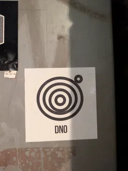 The sticker features a black circular design composed of concentric rings, with a smaller circle offset above and to the right. The acronym 'DNO' is printed below the design.