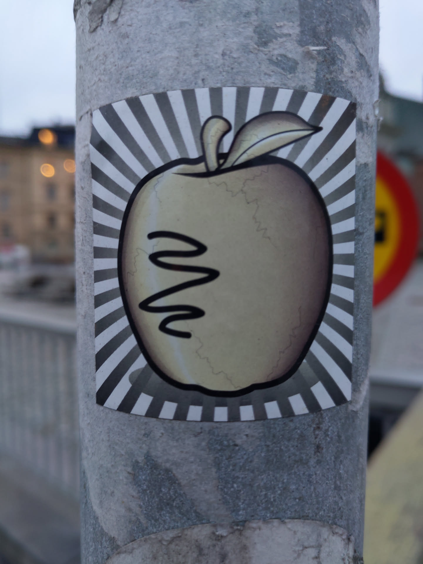 This sticker features a stylized illustration of an apple against a sunburst background.  The apple appears slightly weathered or distressed, adding to its unique charm.  There is a simple design on the apple that resembles a squiggly line or a face. 