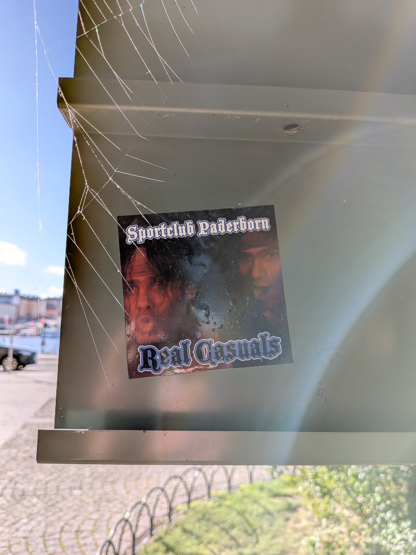 A square sticker placed on a sage-green metal structure, partially covered by fine spiderwebs. The sticker features an image of the pop music duo Milli Vanilli against a dark background. Gothic/blackletter font in white with blue inner shading appears at the top reading 'Sportclub Paderborn' and at the bottom reading 'Real Casuals'.