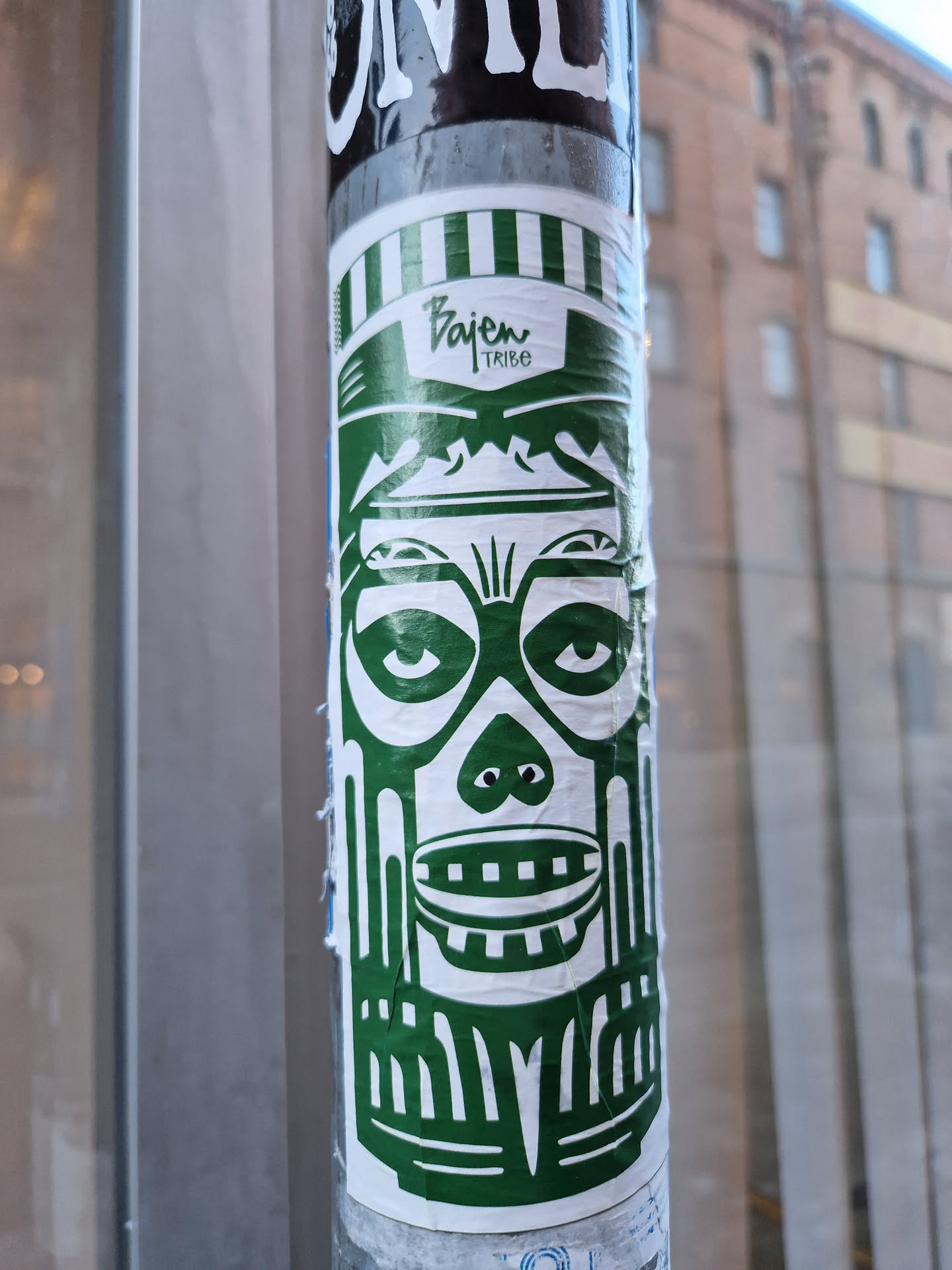 A green and white sticker featuring a stylized tribal mask design. The sticker is rectangular with rounded corners and is applied to a metal pole. The mask has large eyes, a wide mouth, and detailed facial features.  The text "Bajen Tribe" is visible at the top of the sticker.