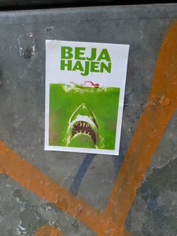 A white sticker affixed to a surface shows an image reminescent of the movie poster of the movie "Jaws", but the text reads "BEJA HAJEN", instead of "JAWS". A swimmer is seen above the surface of a green body of water, while below the water, the shark is about to attack.