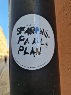 A round white sticker on a black pole with black text that says "Stärning på all plan". The text is handwritten style.