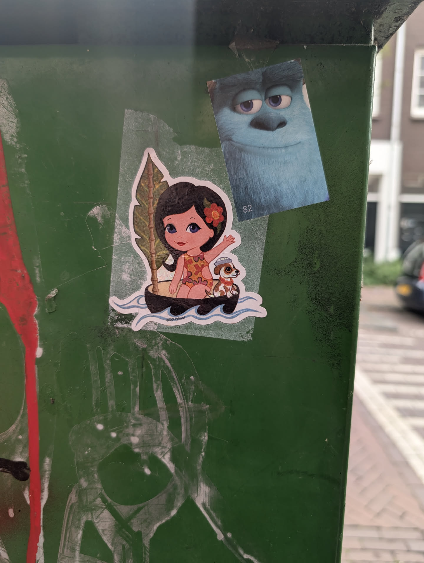 The sticker features a stylized illustration of a young girl with dark hair and a flower behind her ear, wearing a vibrant, cut-out swimsuit. She is seated in a small, dark boat, accompanied by a small dog wearing a sailor's hat. Behind the girl is a large, green tropical leaf. The boat is depicted floating on blue, wavy water. The overall style is cartoony and playful, with bright colors.