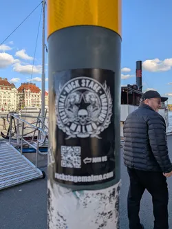 Street sticker A vertical rectangular sticker in black and white stuck to a round metal post. The upper section features a circular emblem with a central star, a skull, wings, and flames, encircled by the text 'FESTIVAL · ROCK STAGE · UNBOUND' and resting on a ribbon displaying 'MALMÖ'. Below this emblem is a QR code alongside an arrow with the prompt 'JOIN US', and at the bottom edge is the website URL 'rockstagemalmo.com'.