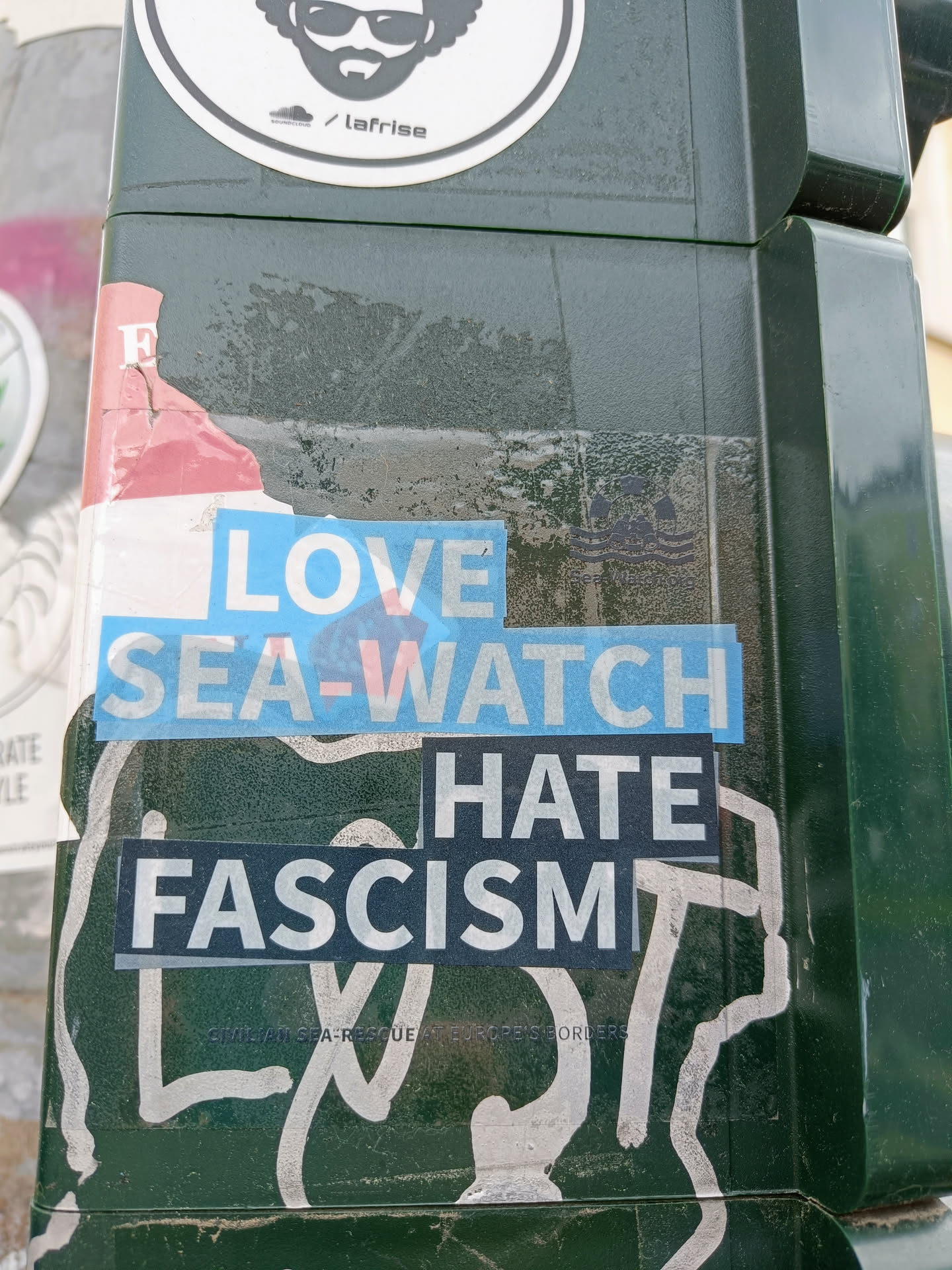 A sticker with the text 'Love Sea-Watch Hate Fascism'. The sticker is placed on a dark green surface, and there are other stickers and graffiti around it.