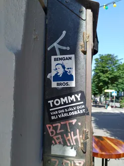 A sticker featuring two portraits and the text 'Bengans Bros.' is affixed to a metal pole.  Below it is another sticker that says 'Tommy' and a Swedish phrase, 'Var dig själv och bli världs bäst'.