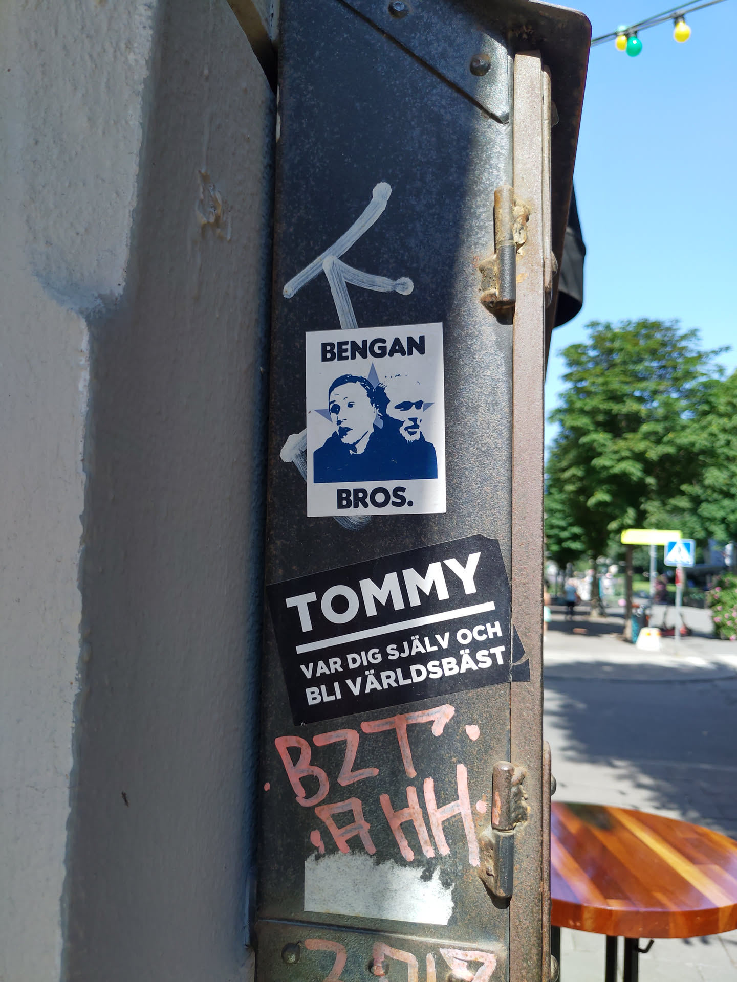 A sticker featuring two portraits and the text 'Bengans Bros.' is affixed to a metal pole.  Below it is another sticker that says 'Tommy' and a Swedish phrase, 'Var dig själv och bli världs bäst'.