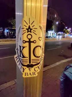 Street sticker A sticker featuring an anchor design with the text "Kitty Cheng Bar" and the initials "KC" inside.  It has a nautical theme with a star, heart, and stylized waves incorporated into the design. The sticker appears to be affixed to a pole outdoors at night.