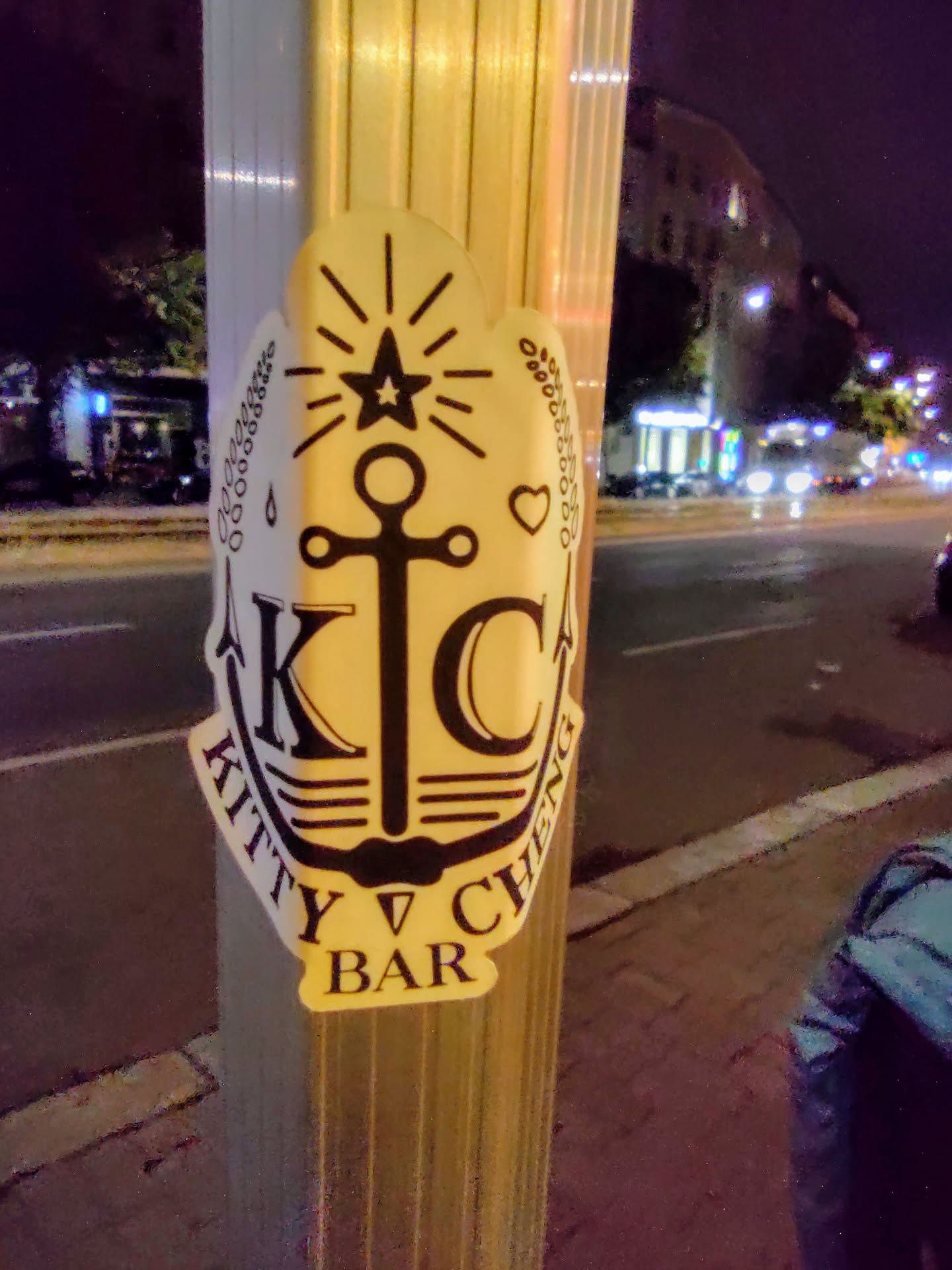 A sticker featuring an anchor design with the text "Kitty Cheng Bar" and the initials "KC" inside.  It has a nautical theme with a star, heart, and stylized waves incorporated into the design. The sticker appears to be affixed to a pole outdoors at night.