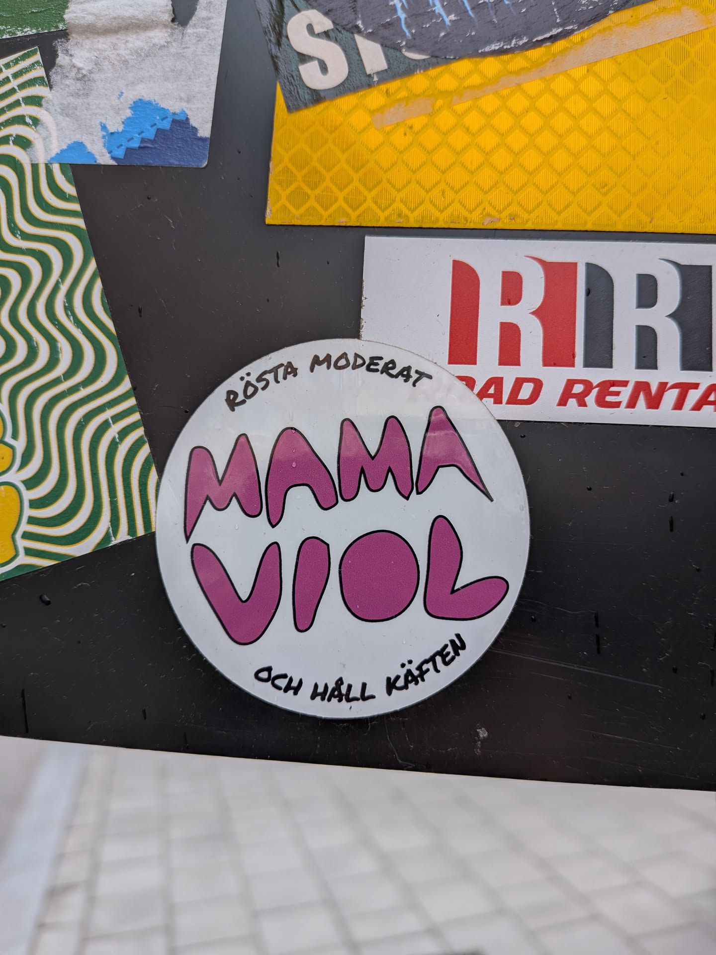 A circular white sticker featuring a central design of the words 'MAMA VIOL' in large, bubbly, pink letters with a black outline. Arched above this central text, in smaller black handwritten-style font, are the words 'RÖSTA MODERAT'. Below the central text, also in the same smaller black handwritten-style font, are the words 'OCH HÅLL KÄFTEN'. The sticker has a slightly worn appearance with some minor scuffs and water droplets visible on its surface.