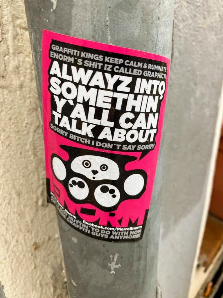 Pink and black sticker with a graphic design and text. The text says "Always into somethin' y'all can talk about." There is also smaller text at the top and bottom. A small cartoon animal is featured on the sticker.  A Facebook link is present at the bottom. 