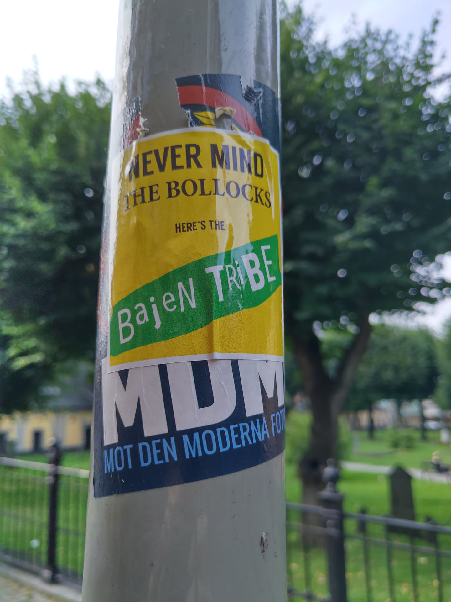 A collage of stickers on a pole. The main sticker is yellow and green with the text "Never mind the bollocks, here's the Bajen Tribe." Below is a dark blue sticker with white text that says "MDM" and "Mot den moderna fotbollen".