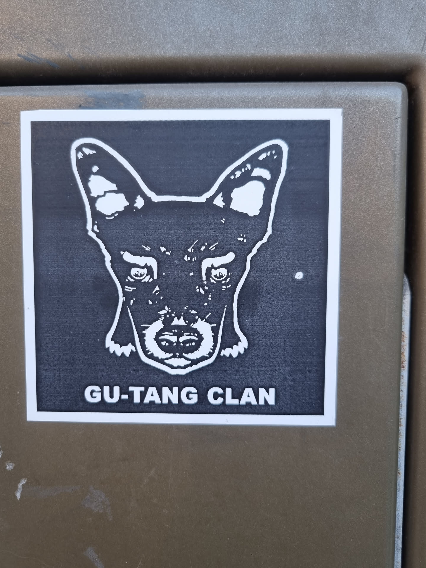 A black and white sticker with a stylized image of a dog's head and the text "GU-TANG CLAN" at the bottom. The dog's expression is serious and intense. The sticker has a simple design, suggesting a possible affiliation with a group or brand.