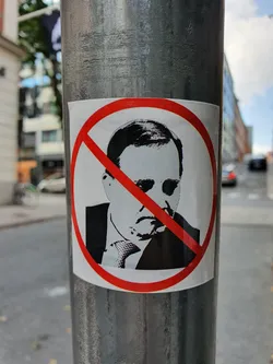 A sticker depicting a prominent figure with a red circle and cross symbol, indicating disapproval or rejection. The sticker is affixed to a metal pole in an urban setting.