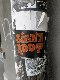 A black sticker with the text 'SIENA ROOT' written in a retro, orange and yellow font. The sticker has a slightly worn look and is affixed to a weathered surface, suggesting it has been there for some time.