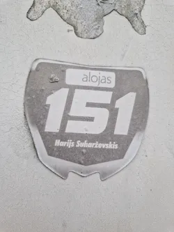 A gray and white sticker with the number 151, the word alojas and the name Harjis Suharževskis. The sticker has a slightly worn look.