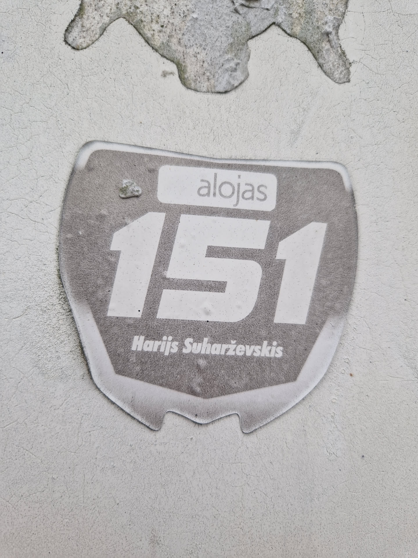 A gray and white sticker with the number 151, the word alojas and the name Harjis Suharževskis. The sticker has a slightly worn look.