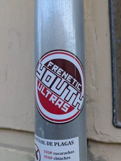 A round sticker with the words "Frenetic Youth Ultras" on a metal pole. The sticker has a white border, a red ring, and a maroon circle in the center. The words "Frenetic" and "Ultras" are in a red font, while the word "Youth" is in a white block font.