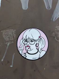 The sticker features an illustration of a girl with glasses and pigtails, licking a pink lollipop. The artwork is done in a white and pink color scheme with black line art. The girl has freckles, a choker, and is depicted with a playful, somewhat mischievous expression.