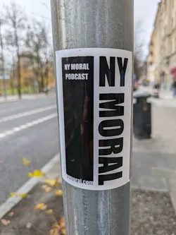 A black and white sticker advertising the NY Moral Podcast. The sticker is affixed to a metal pole.  The text on the sticker reads 'NY Moral Podcast' and 'nymora.com'.