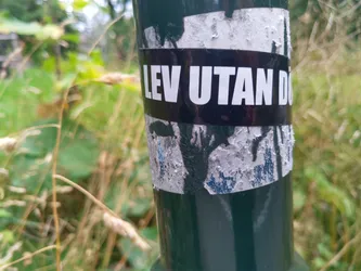 A sticker on a green pole with the text "LEV UTAN DOGMA" in white letters on a black background. The sticker is partially damaged and peeling. The background is blurred and shows green vegetation.