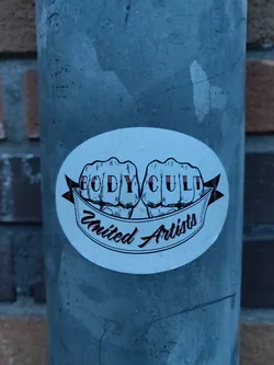 A round sticker featuring two fists with the words BODY CULT in between them, and the words "United Artists" on a banner below. The design is in black and white with a hint of red.
