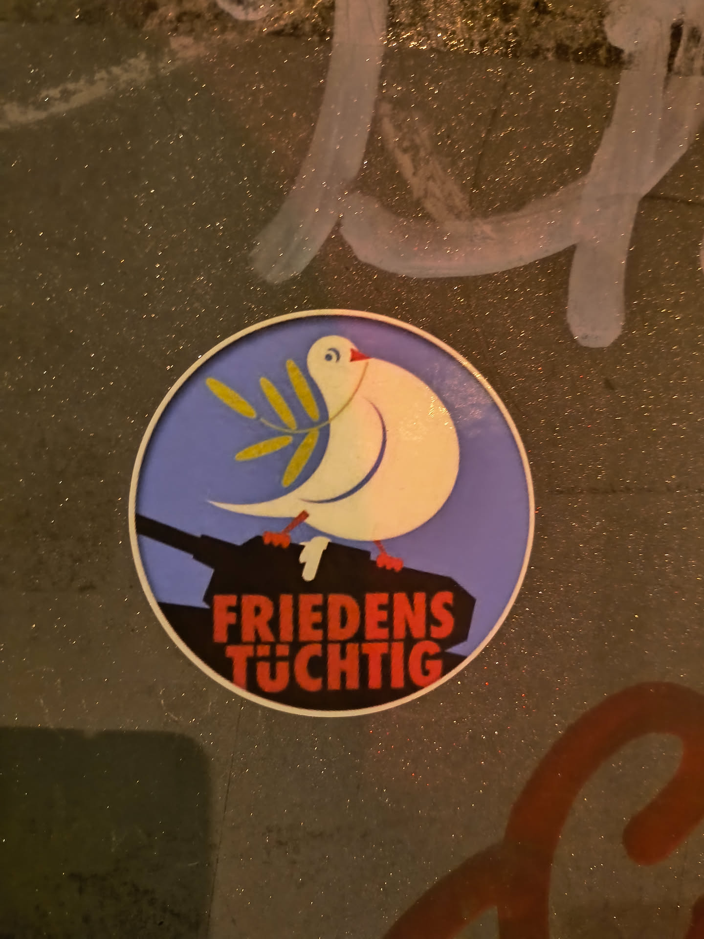 A round sticker featuring a white dove carrying an olive branch, perched atop a tank. The text below reads "FRIEDENS TÜHTIG" which translates from German to "Peace-capable". The design promotes peace and disarmament.