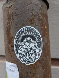 A circular sticker predominantly in black, white, and shades of grey, featuring a stylized, somewhat distressed or contemplative face with spiky hair. The figure's hands are positioned near its ears. Above the head, text arches in a decorative font, and below the face, within a circular, globe-like element, more text is present. The background behind the figure includes intricate, swirling patterns, and the bottom portion of the sticker features a black and white checkerboard pattern. The entire design is outlined with a clean white border.