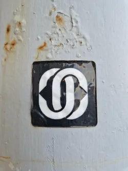 Close-up of a square sticker affixed to a weathered surface. The sticker features a bold, interlocking design in white against a black background. The surface shows signs of rust and wear.