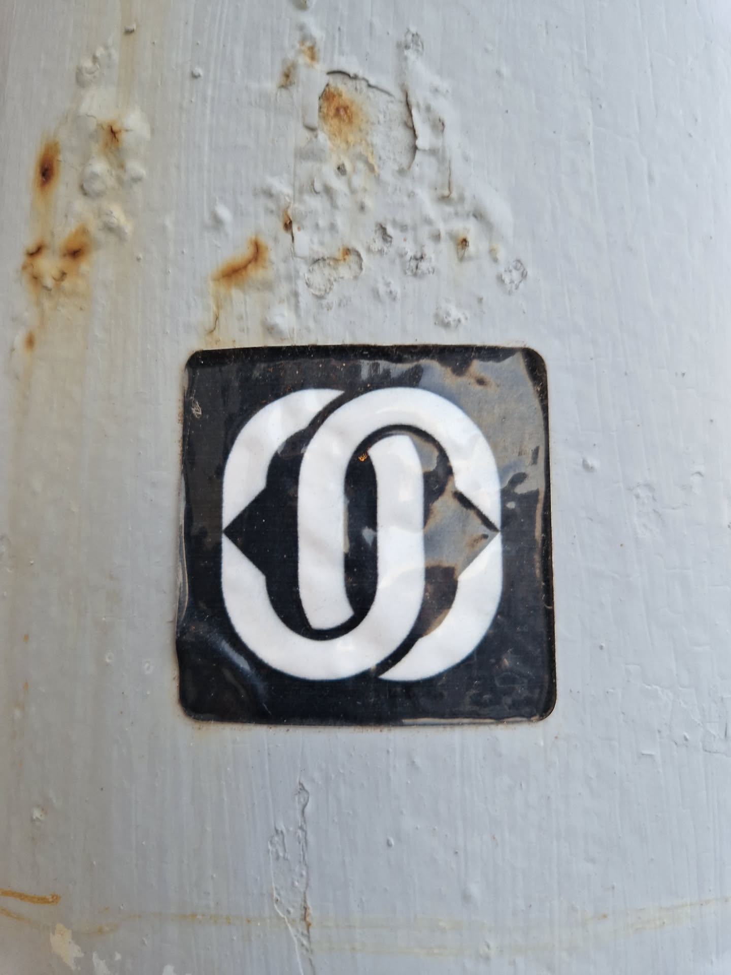 Close-up of a square sticker affixed to a weathered surface. The sticker features a bold, interlocking design in white against a black background. The surface shows signs of rust and wear.