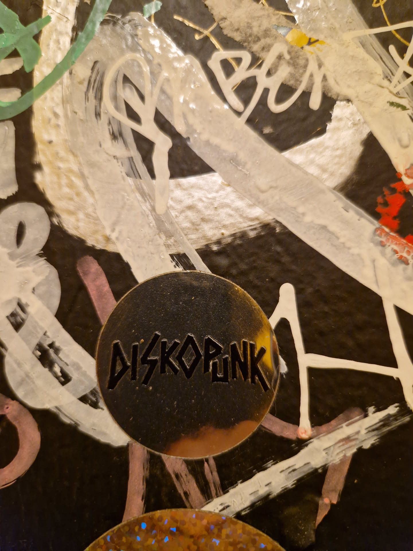 A round, metallic gold sticker with the word "DISKOPUNK" in a bold, black, stylized font. It is affixed to a wall covered in various graffiti tags and paint splatters.