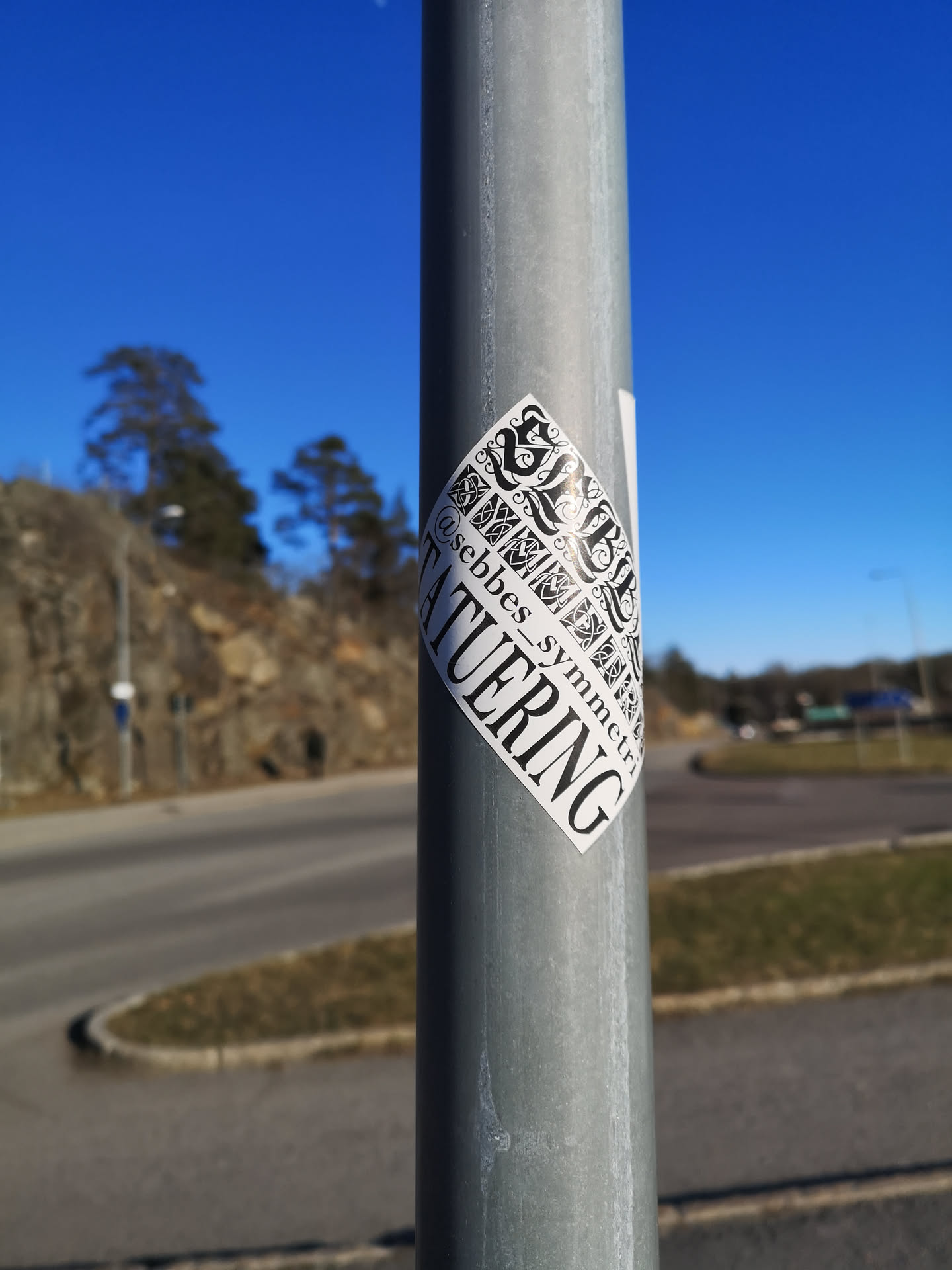 A black and white sticker with a distinctive vertical diamond or leaf shape, featuring intricate, symmetrical knotwork patterns in the upper two-thirds. The design is reminiscent of Celtic or Norse art, with swirling, interlocking lines. The lower third contains text: '@sebbes_symmetry' is written horizontally along the left curve, and 'TATUERING' is written vertically in large, bold letters along the right curve. The sticker has a clean, graphic aesthetic.