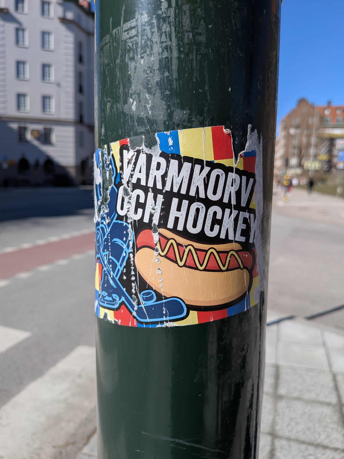 The sticker features a vibrant, cartoon-style illustration centered around a hot dog with mustard in a bun. To the left of the hot dog, a blue hockey stick and puck are depicted. The background is a colorful, distressed pattern of vertical and horizontal stripes in red, yellow, and blue, reminiscent of the Swedish flag. Above the hot dog, white block letters on a black background clearly spell out 'VÄRMKORV OCH HOCKEY'. The sticker is rectangular but shows significant wear, with tears and peeling along its edges, revealing the dark green pole it's affixed to.