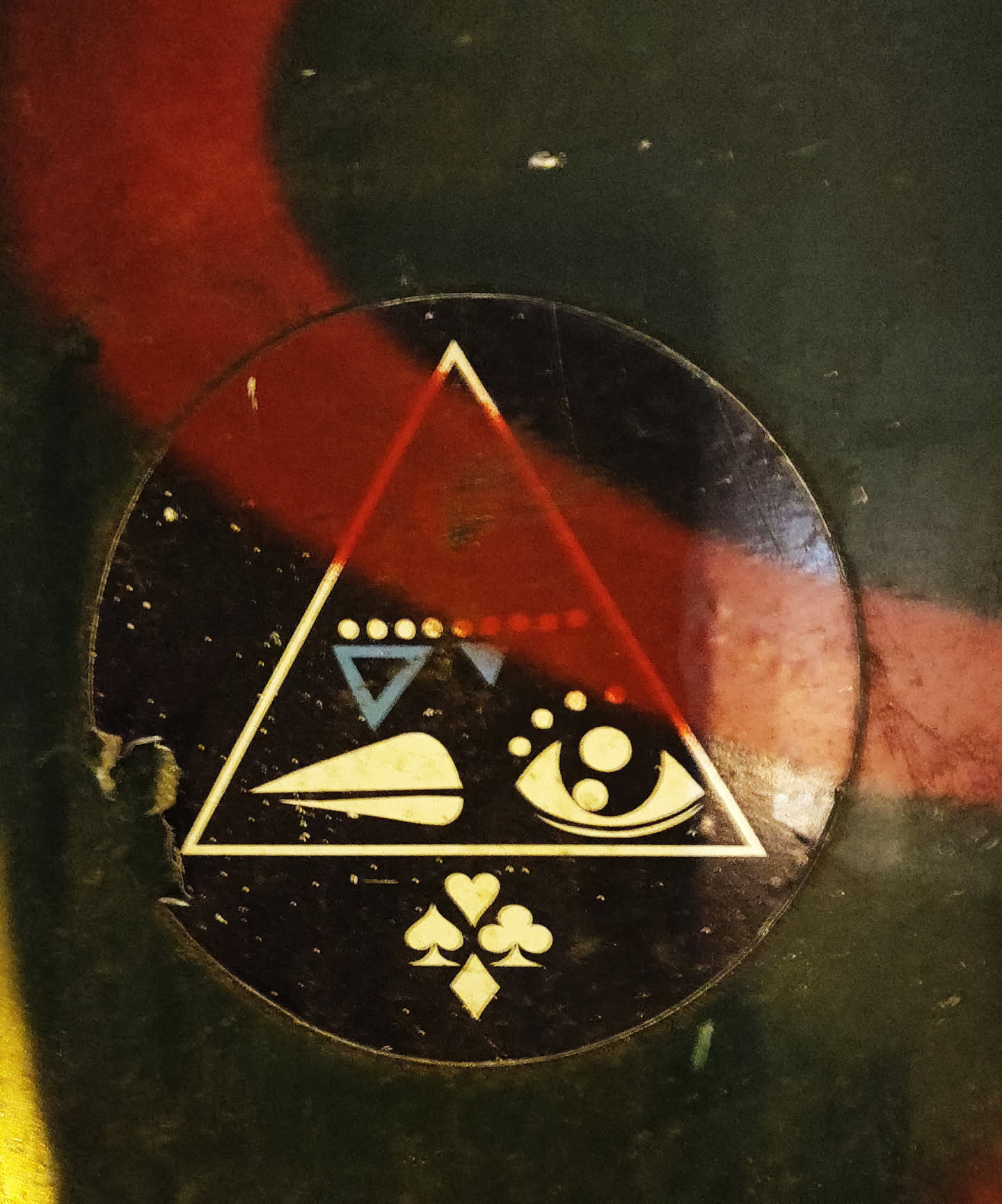 A round sticker featuring a red triangle with an eye, symbols, and playing card suits at the bottom. The style is mystical and symbolic. The sticker shows some wear.