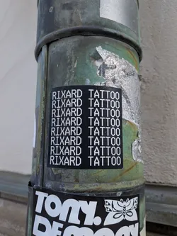 A rectangular black sticker adhered to a weathered green pipe. The sticker features the phrase 'RIXARD TATTOO' displayed in all-caps using a white, pixelated, 8-bit style typeface, repeated vertically down the sticker in eight uniform rows.