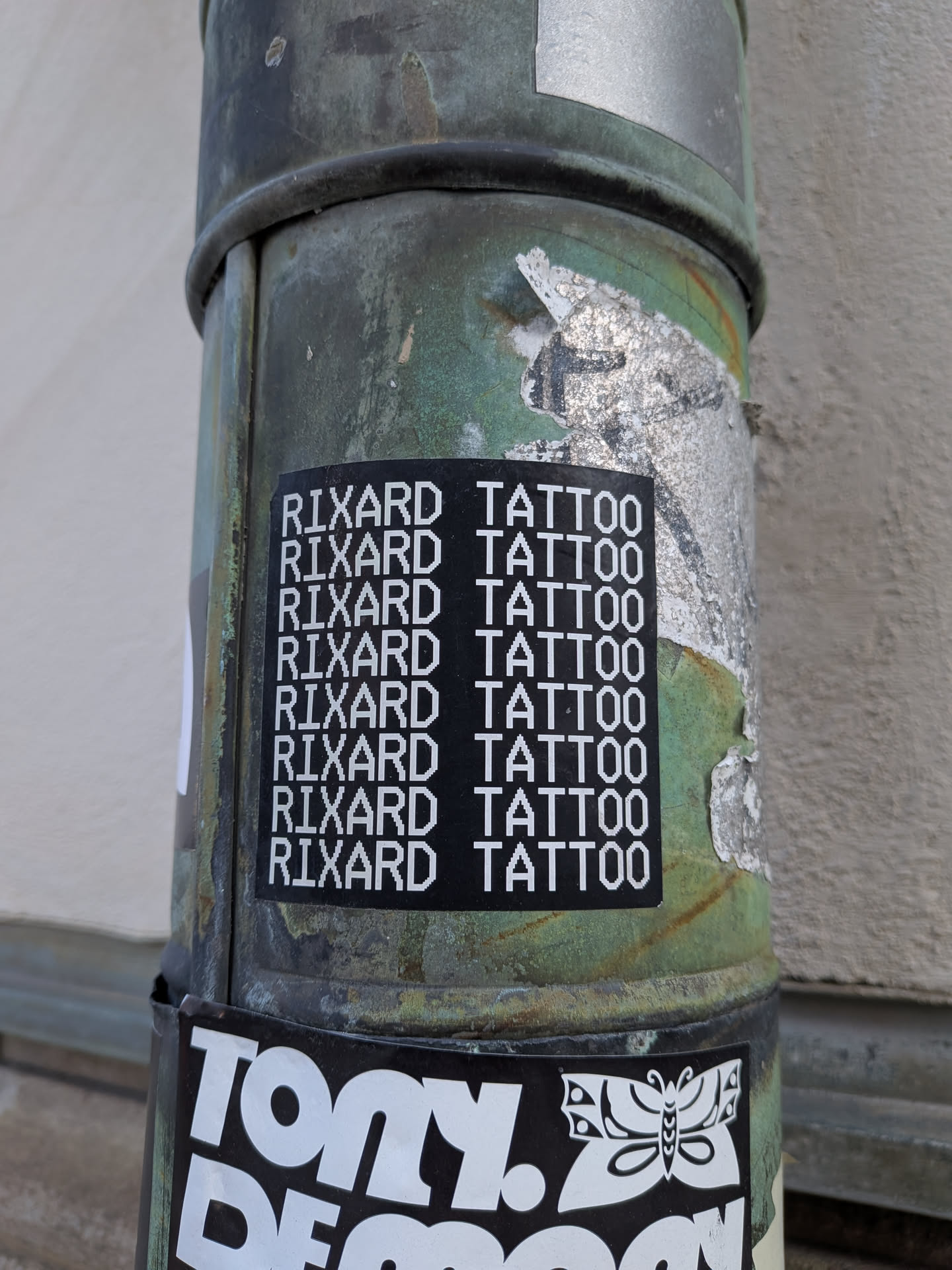 A rectangular black sticker adhered to a weathered green pipe. The sticker features the phrase 'RIXARD TATTOO' displayed in all-caps using a white, pixelated, 8-bit style typeface, repeated vertically down the sticker in eight uniform rows.