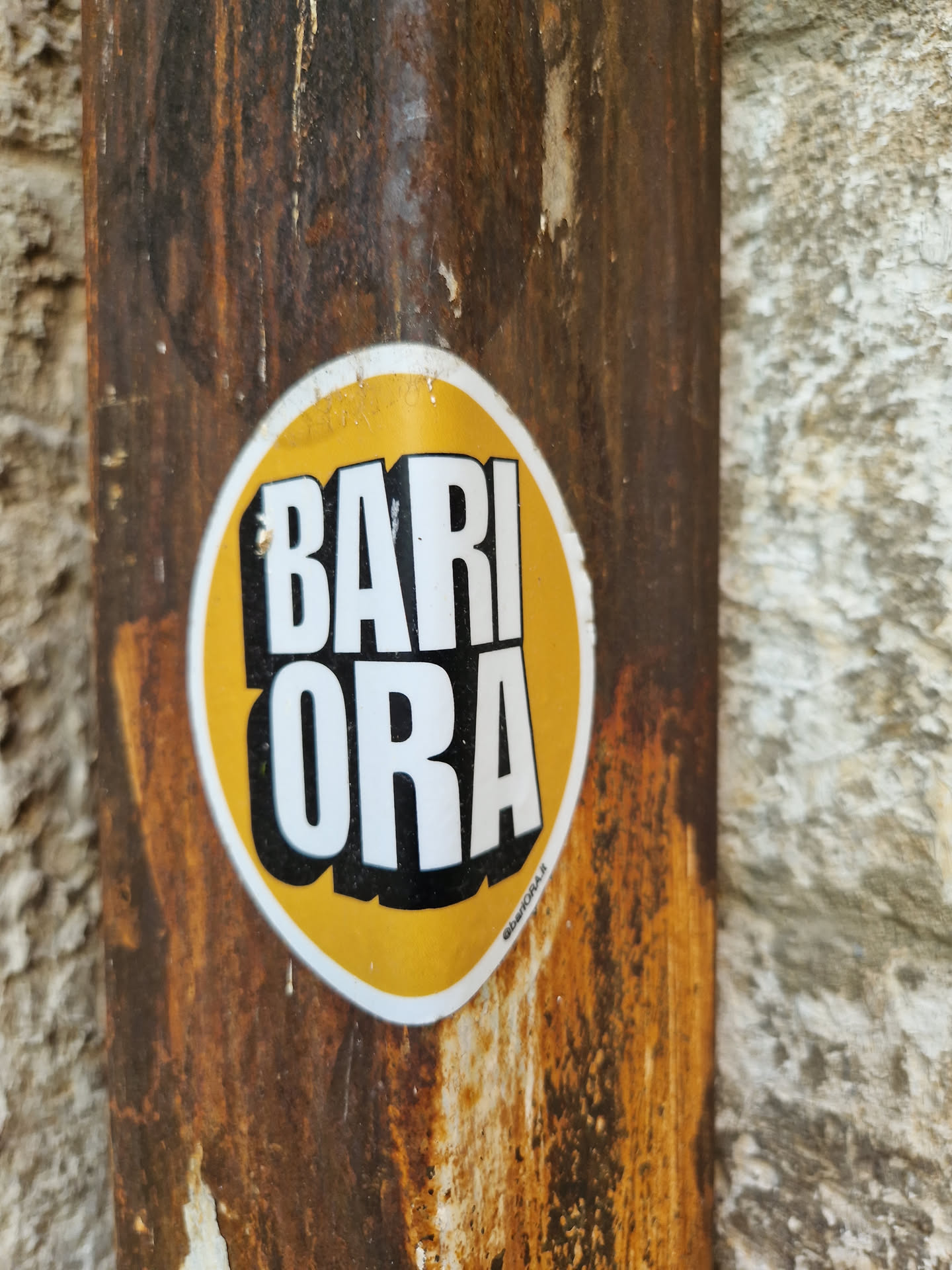 A yellow and black sticker with the words "BARI ORA" in a bold, stylized font. The sticker is affixed to a rusty metal surface, suggesting it may be street art or urban decor. The overall aesthetic is slightly grungy, enhancing its visual appeal.