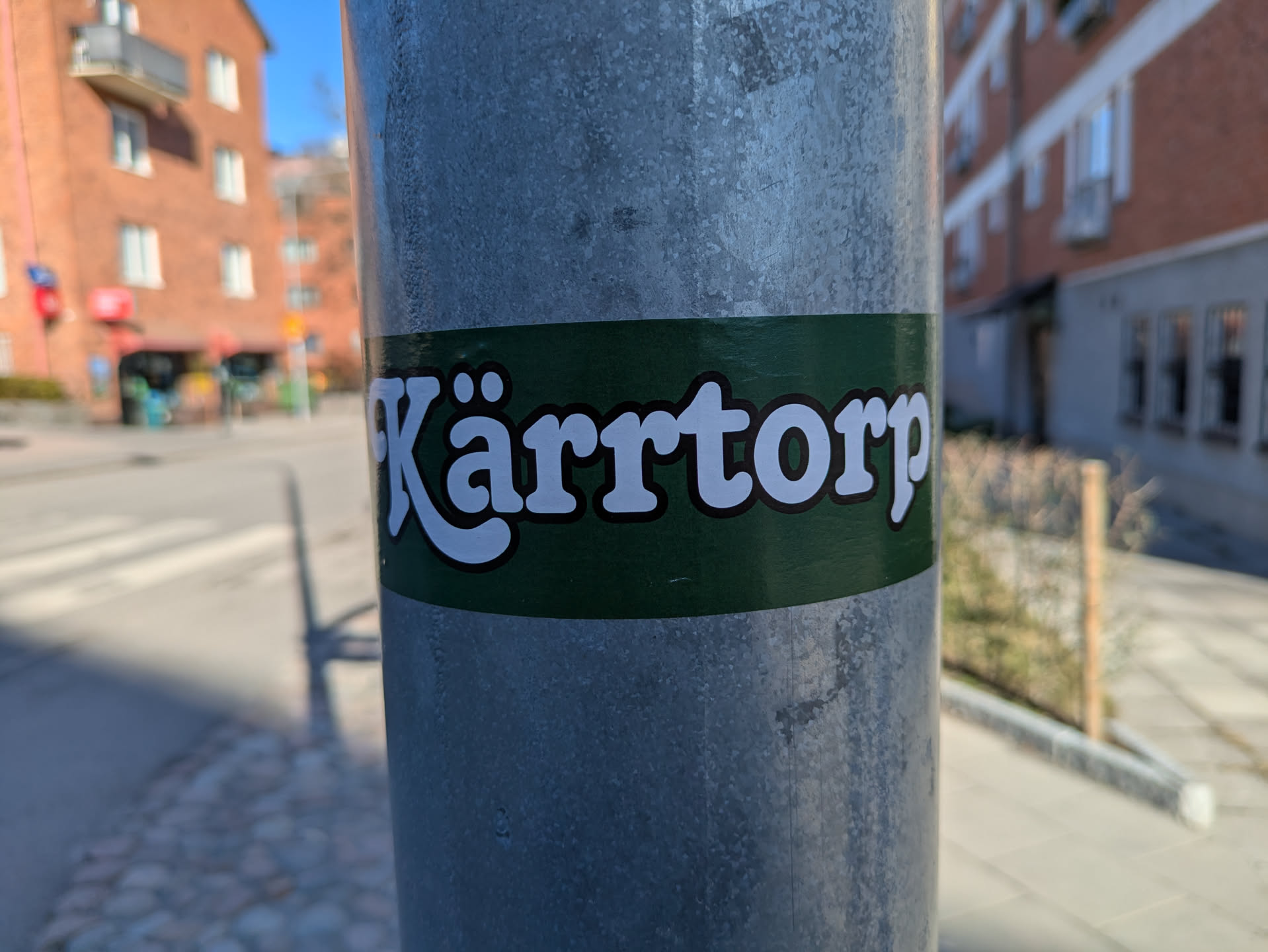 A rectangular sticker with a dark green background and the text 'Kärrtorp' in white with a brown outline, placed on a metal pole.