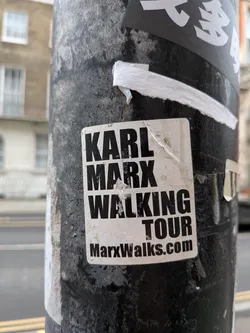 A rectangular sticker with slightly rounded corners, featuring bold black text on a white background. The text is arranged in a block format, reading 'KARL MARX' on the first line, 'WALKING' on the second, and 'TOUR' on the third. Below this, in a smaller font, is 'MarxWalks.com'. The sticker shows signs of wear, including dirt, scuff marks, and slight peeling at the edges, particularly the top right corner, indicating it has been exposed to the elements on a textured, dark pole.