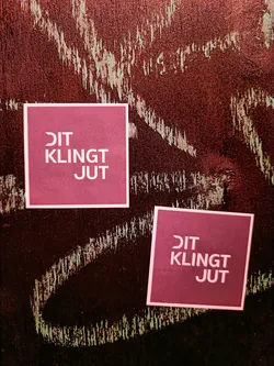 Two square stickers on a dark red surface.  The stickers are maroon with light pink text that says "DIT KLINKT JUT".