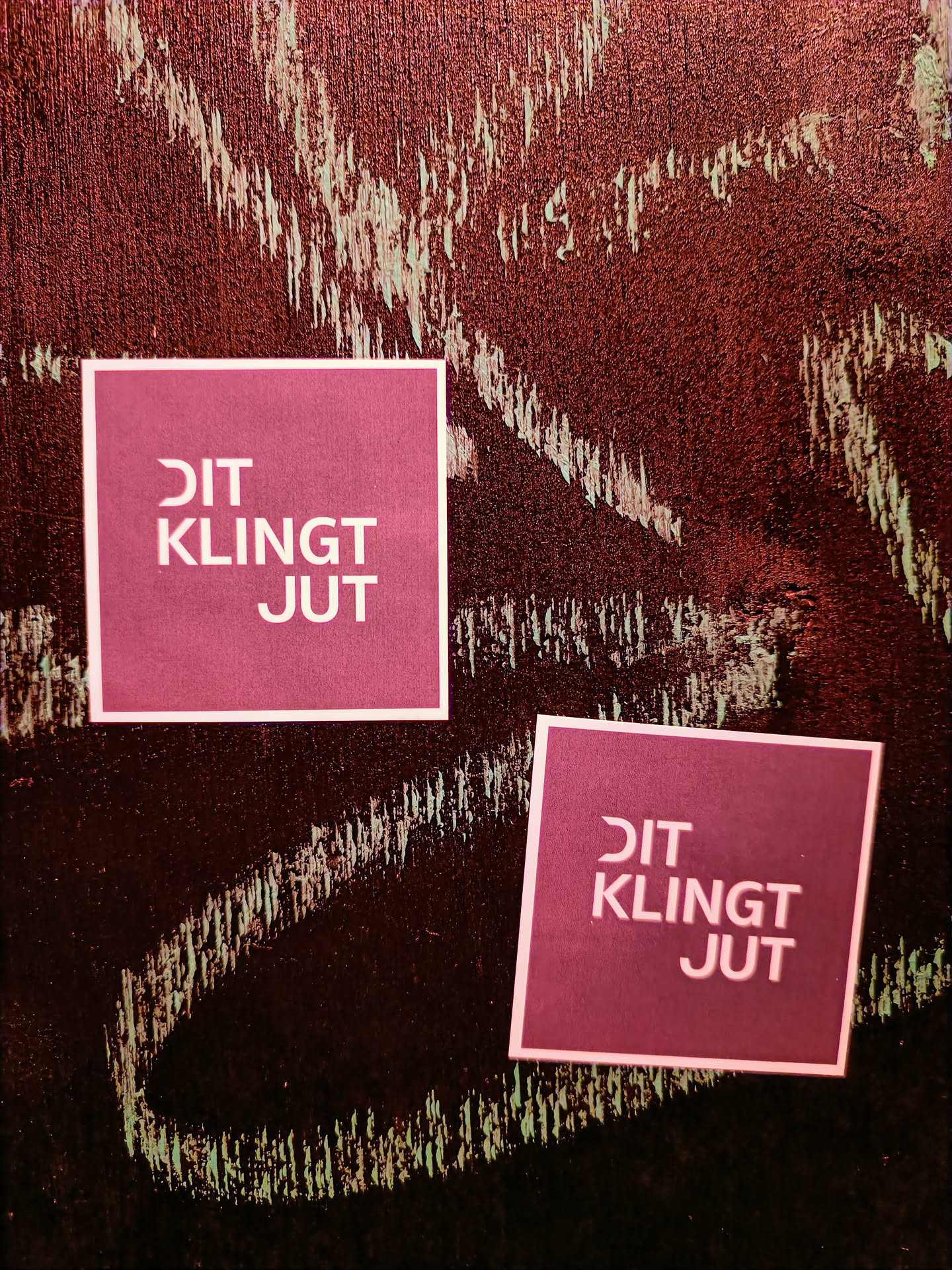 Two square stickers on a dark red surface.  The stickers are maroon with light pink text that says "DIT KLINKT JUT".